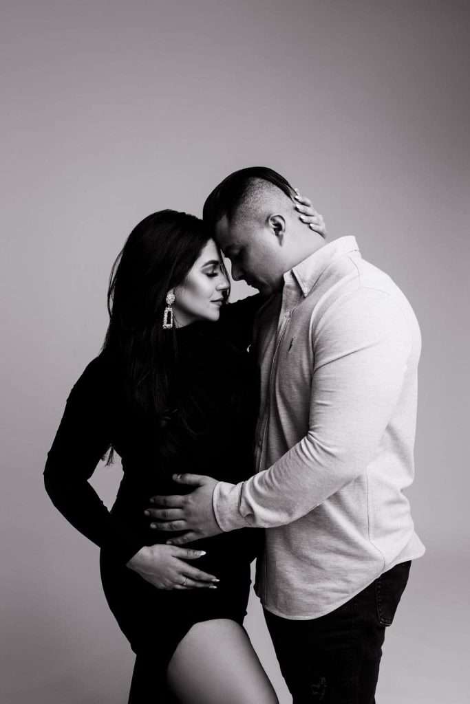 A couple stands close together in an intimate pose, with the man's hand on the woman's waist and their foreheads touching, against a plain background.