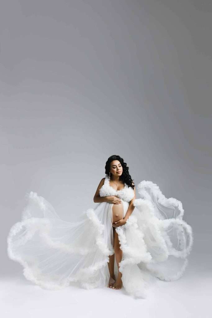 A pregnant woman in a flowing, white, sheer gown stands barefoot against a plain gray background, holding her belly and looking down.