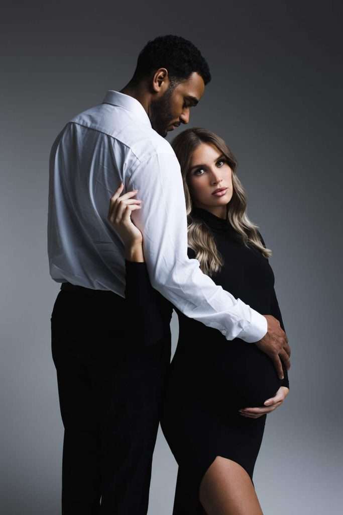 A man in a white shirt stands beside a pregnant woman in a black dress, with his hand on her belly. Both look toward the camera against a neutral background.