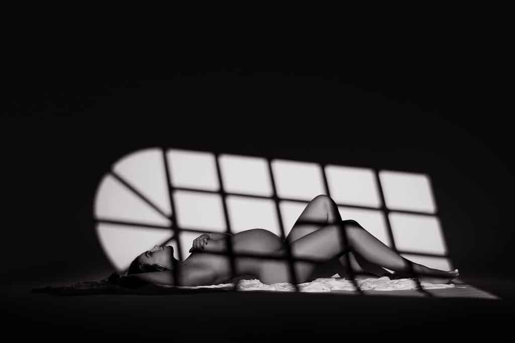 A pregnant woman lies on her side on a blanket, with shadows from a window pattern cast across her body in a dark room.
