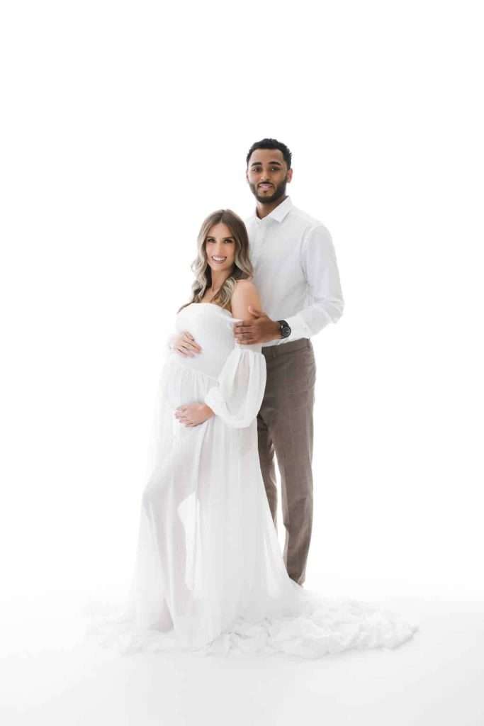 A pregnant woman in a white dress stands in front of a man in a white shirt and beige pants; both are smiling and facing the camera against a white background.