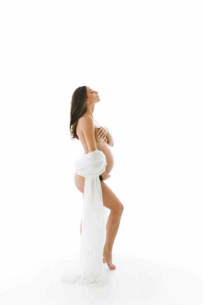Pregnant woman standing sideways, draped in a white fabric, looking upward against a white background.