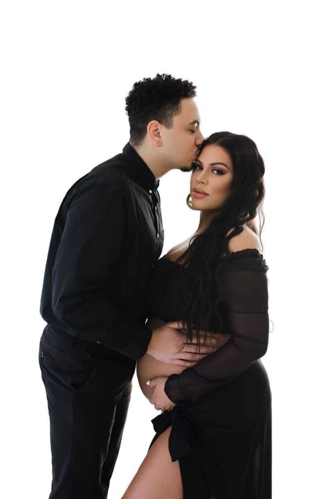 A man in black kisses a woman on the forehead while holding her baby bump; the woman, also in black, stands beside him with one leg showing, both posing against a white background.