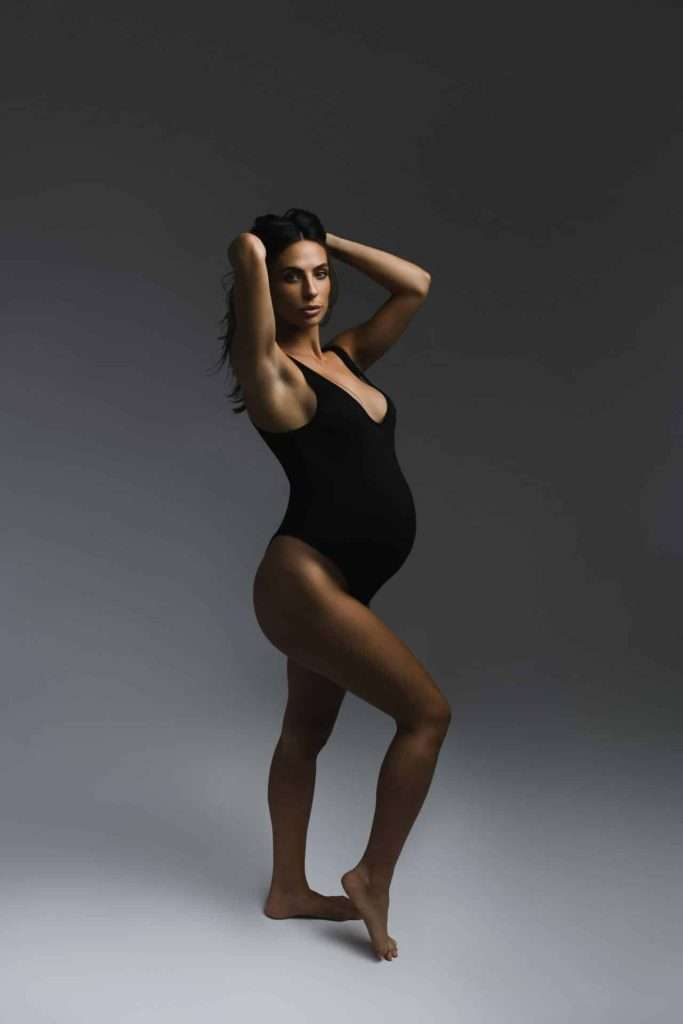 A pregnant woman in a black bodysuit stands barefoot with her hands behind her head against a dark, neutral background.