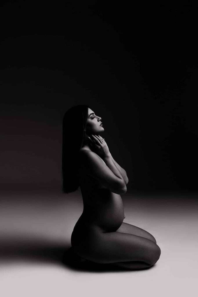 A pregnant woman kneels on the floor in a dark, studio-lit setting, with her hands gently touching her neck and her head slightly tilted back.
