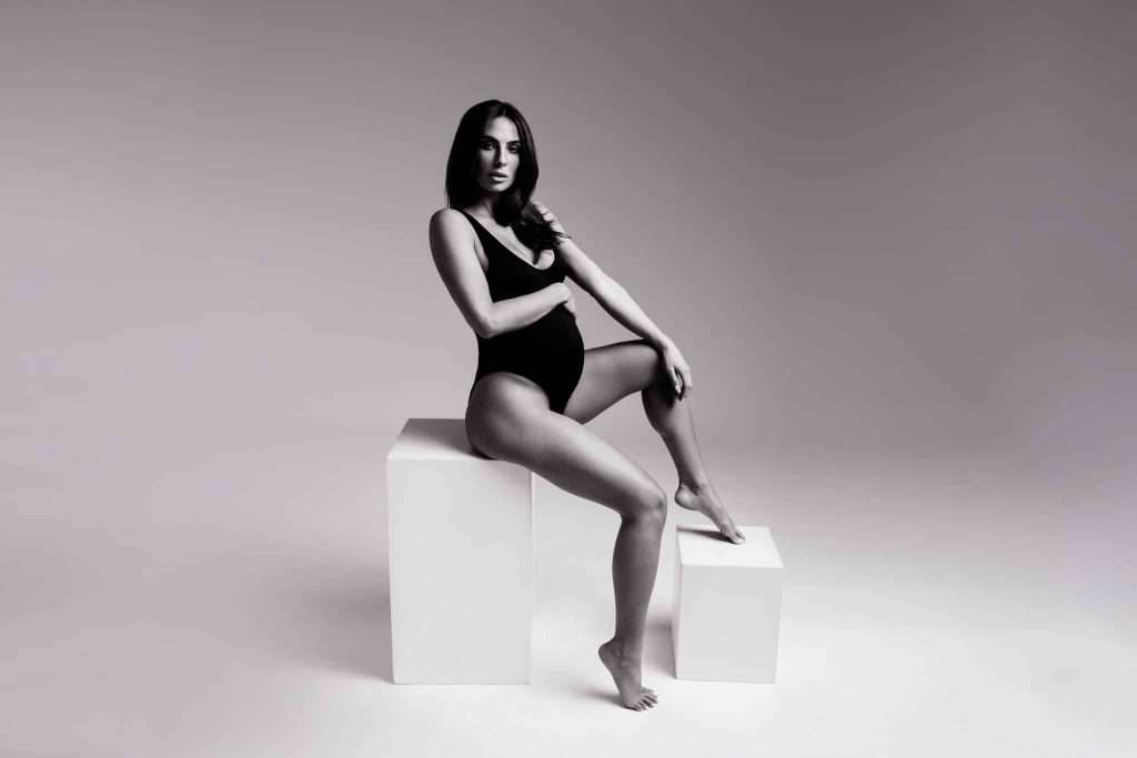 A pregnant woman wearing a black bodysuit sits on a white block with one leg bent and her arm resting on her knee in a studio with a plain background.