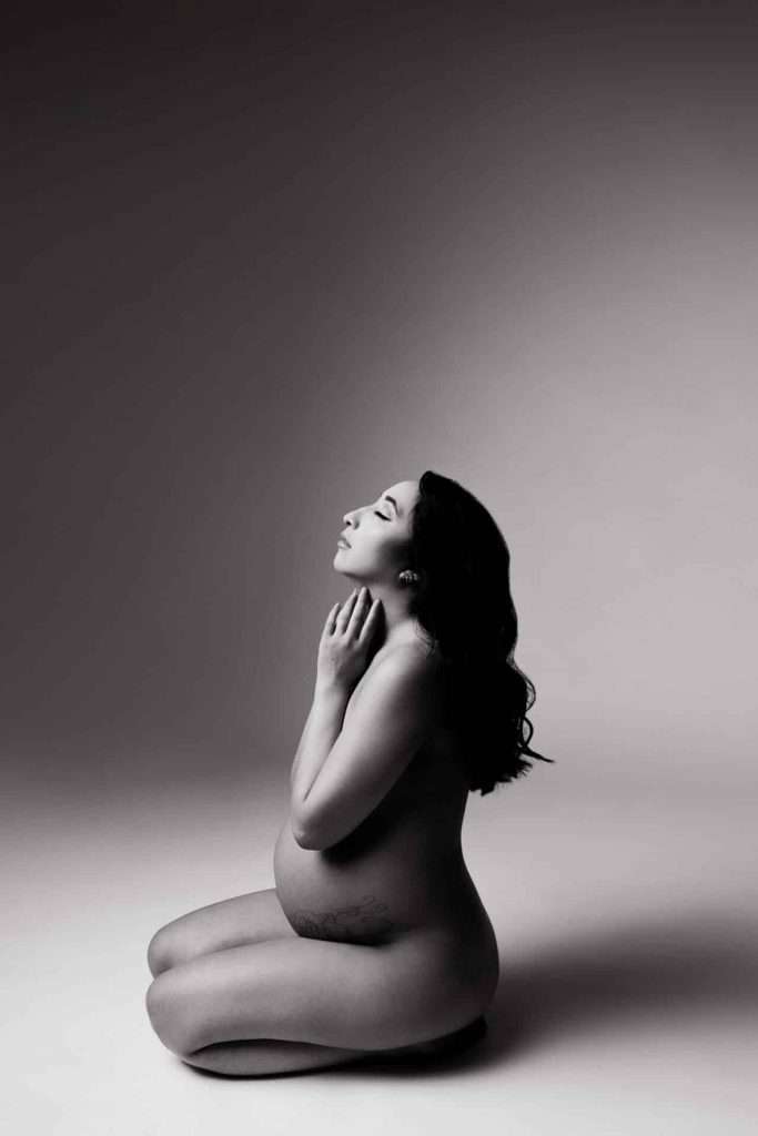 A pregnant woman kneels on the floor in a studio setting, eyes closed and hands near her face, with soft lighting and a neutral background.