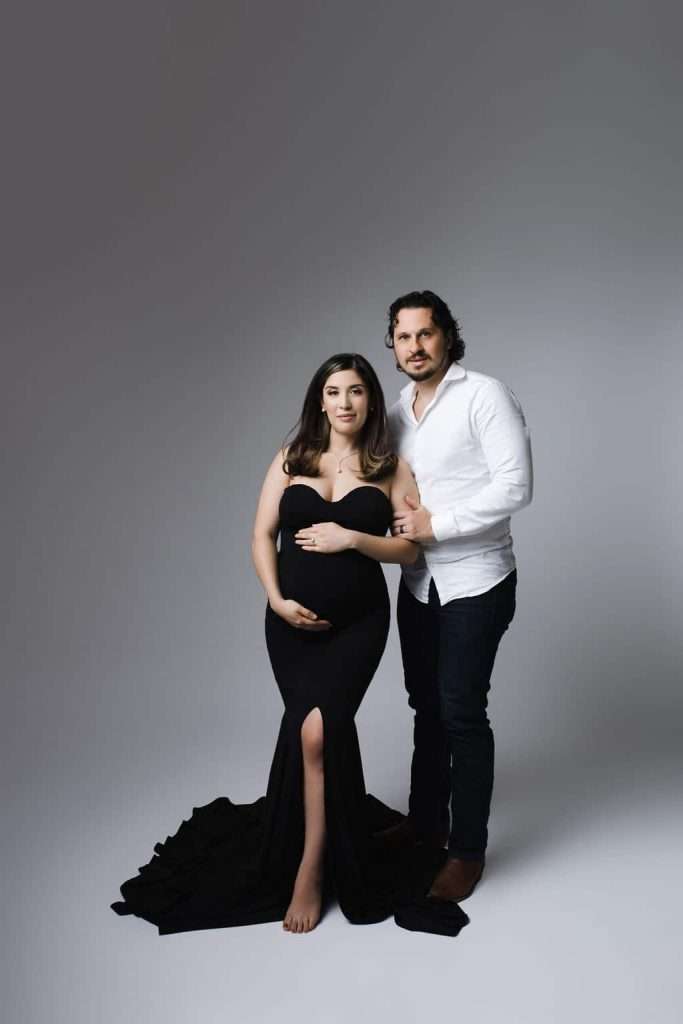 A pregnant woman in a black dress stands beside a man in a white shirt and jeans, both looking at the camera against a plain gray background.