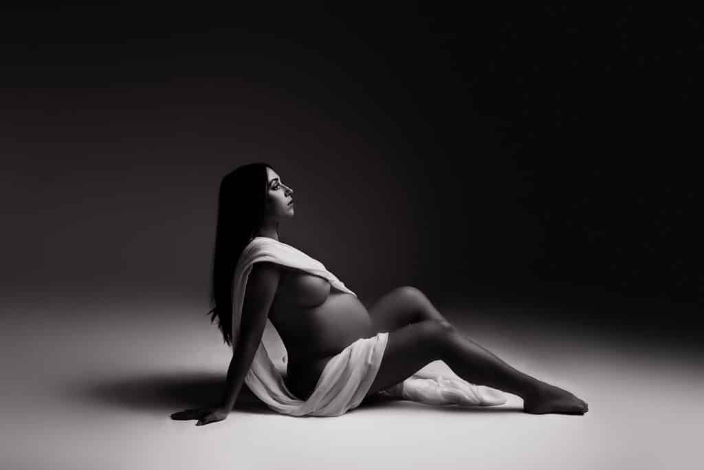 A pregnant woman draped in a white cloth sits on the floor in a studio setting, looking upward; the image is in black and white.