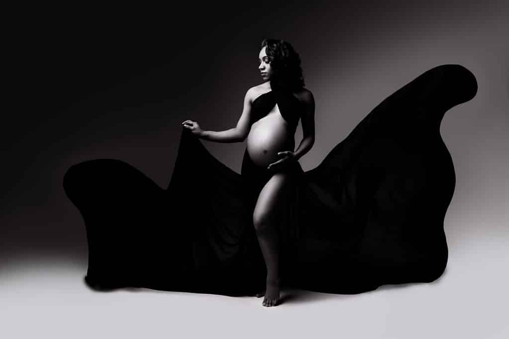 Pregnant woman stands barefoot, draped in flowing black fabric, holding part of the cloth as it billows around her in a studio setting with dramatic lighting.
