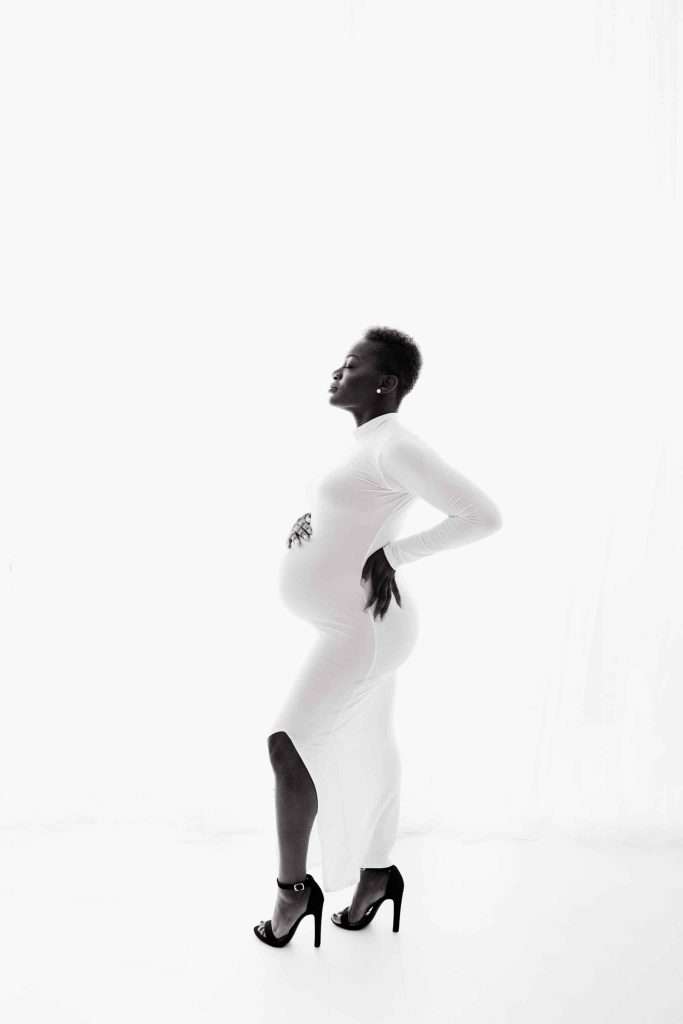 A pregnant woman in a long white dress and high heels stands in profile against a white background, with one hand on her back and the other on her belly.