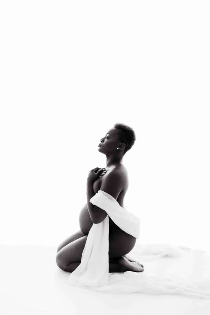 A pregnant person kneels on the floor with eyes closed, draped in a white cloth against a white background.