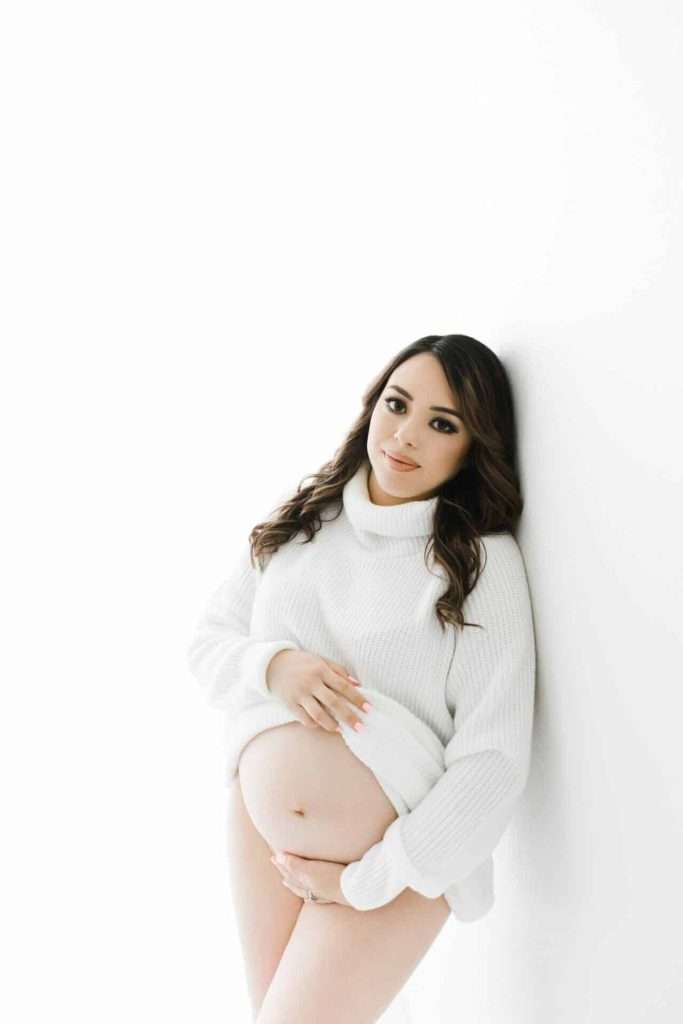 A pregnant woman in a white sweater leans against a white wall, holding her belly and looking at the camera.