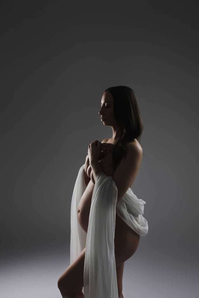 A pregnant woman stands in profile against a gray background, draped in sheer white fabric, holding her chest with one arm and her belly with the other.