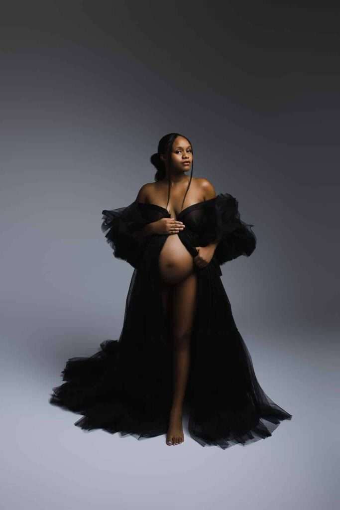 A pregnant woman in a sheer black gown stands barefoot against a plain, softly lit background, looking to the side with one hand on her belly.