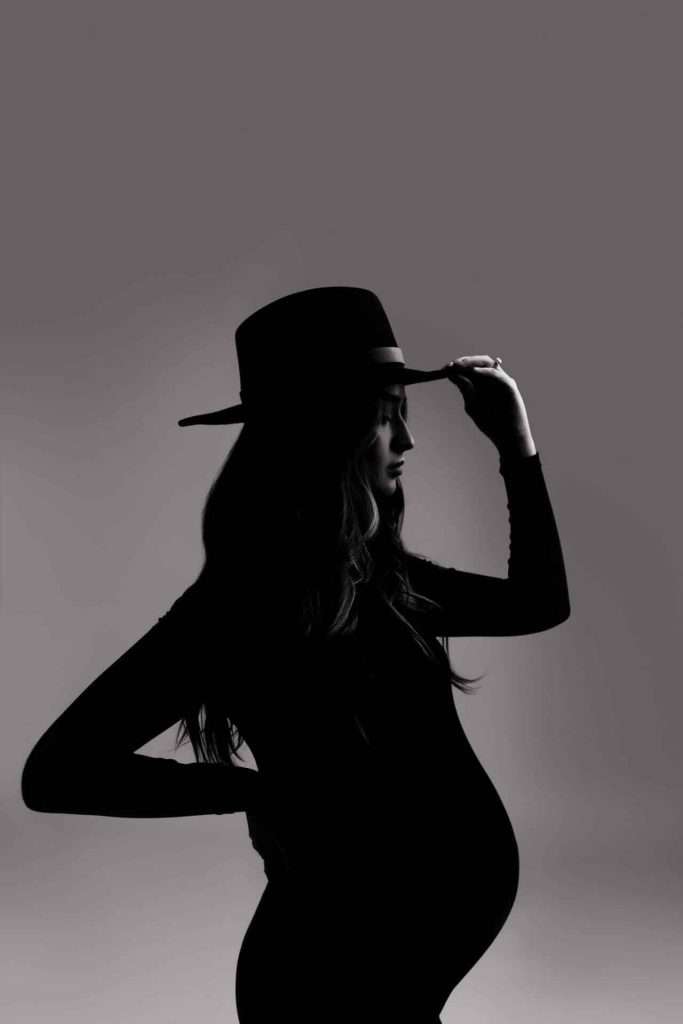 Silhouette of a pregnant woman wearing a hat, standing sideways with one hand on her hip and the other adjusting her hat.