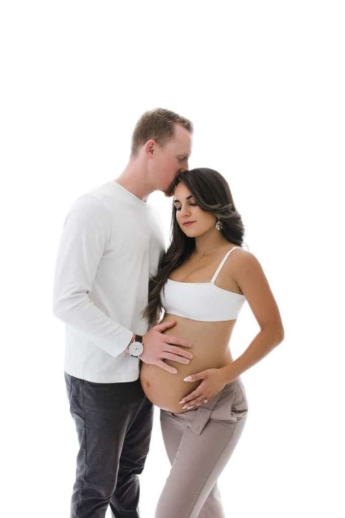 A man kisses the forehead of a pregnant woman while gently holding her belly. Both are standing against a white background.