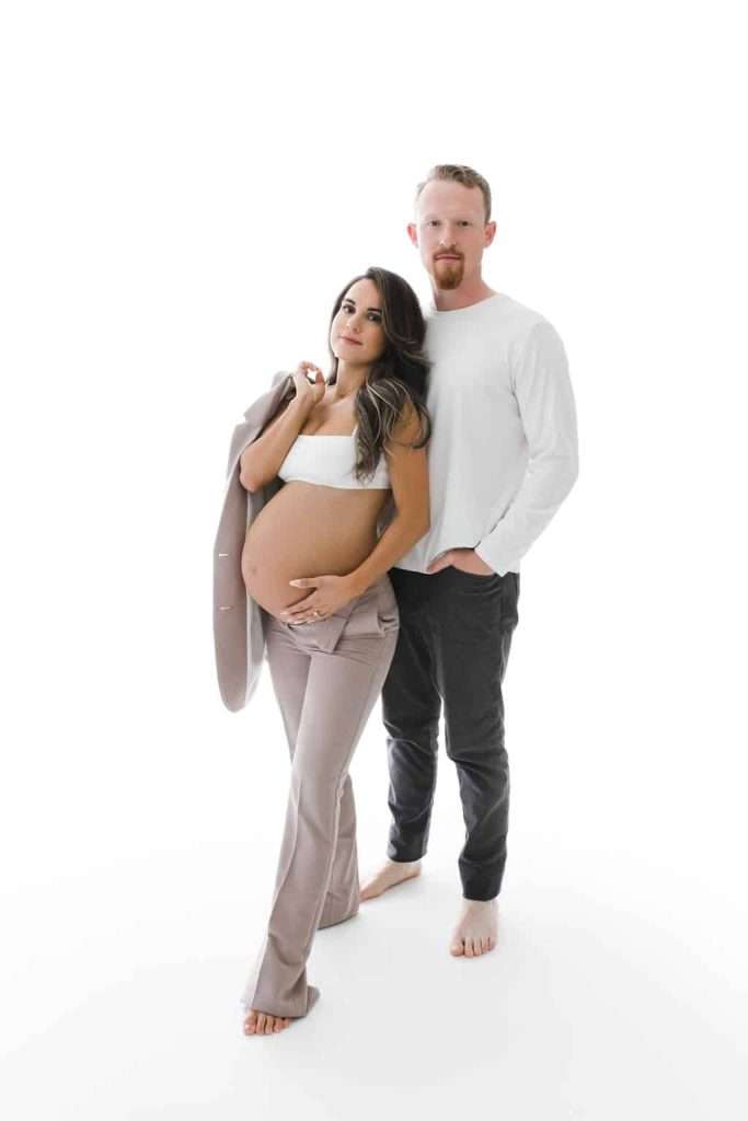 Pregnant woman in a white top and beige pants stands next to a man in a white shirt and black pants, both barefoot, against a white background.