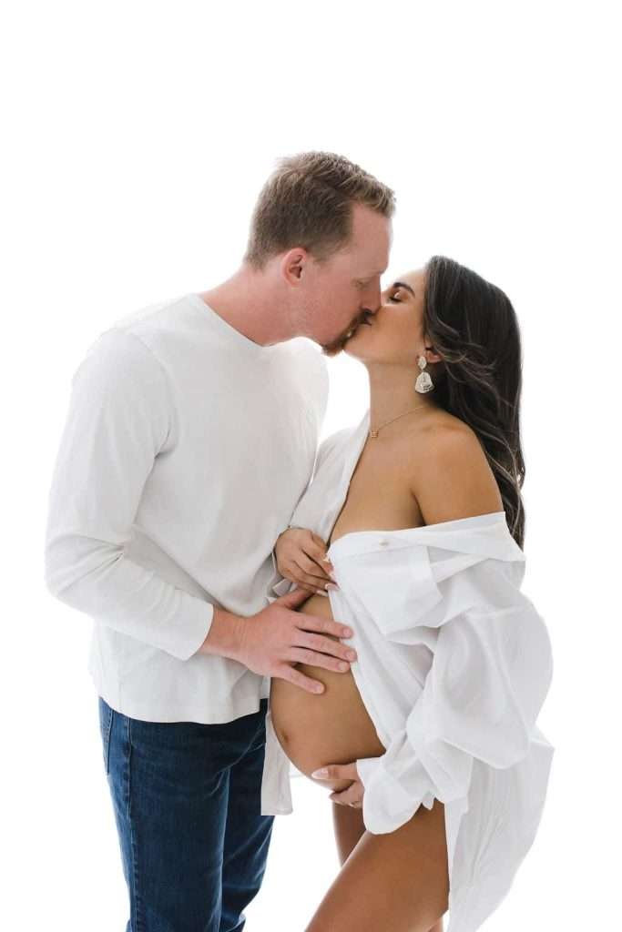 A man and a pregnant woman kiss while standing. The woman is wearing an open white shirt, revealing her baby bump. The man rests his hand on her belly.