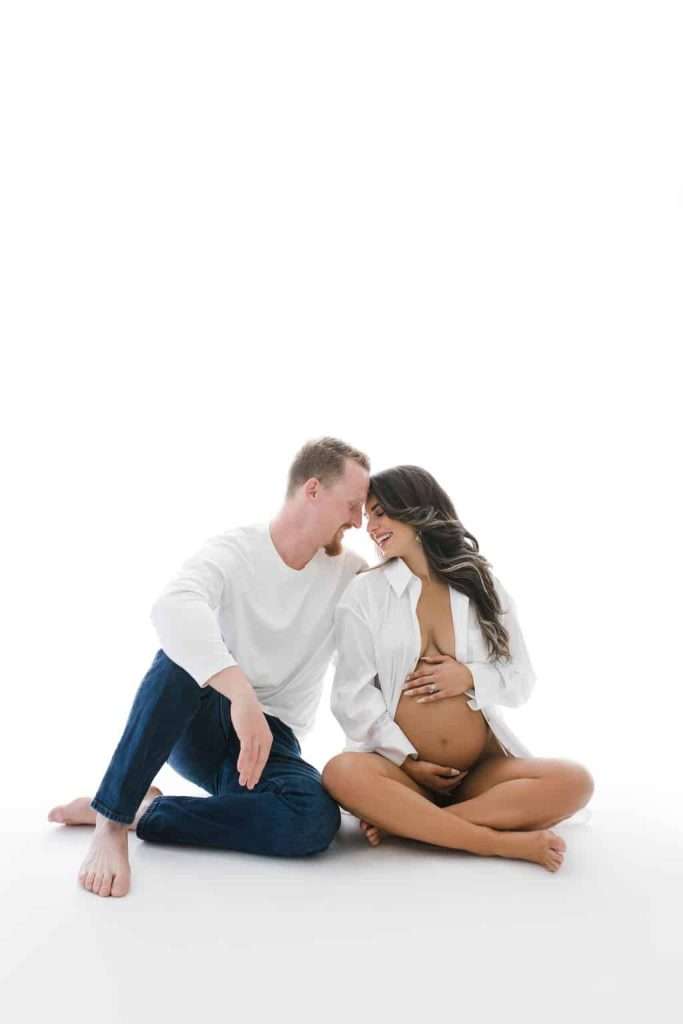 A couple sits on the floor against a white background; the woman is pregnant and smiling, with one hand on her belly, while the man leans in close to her.