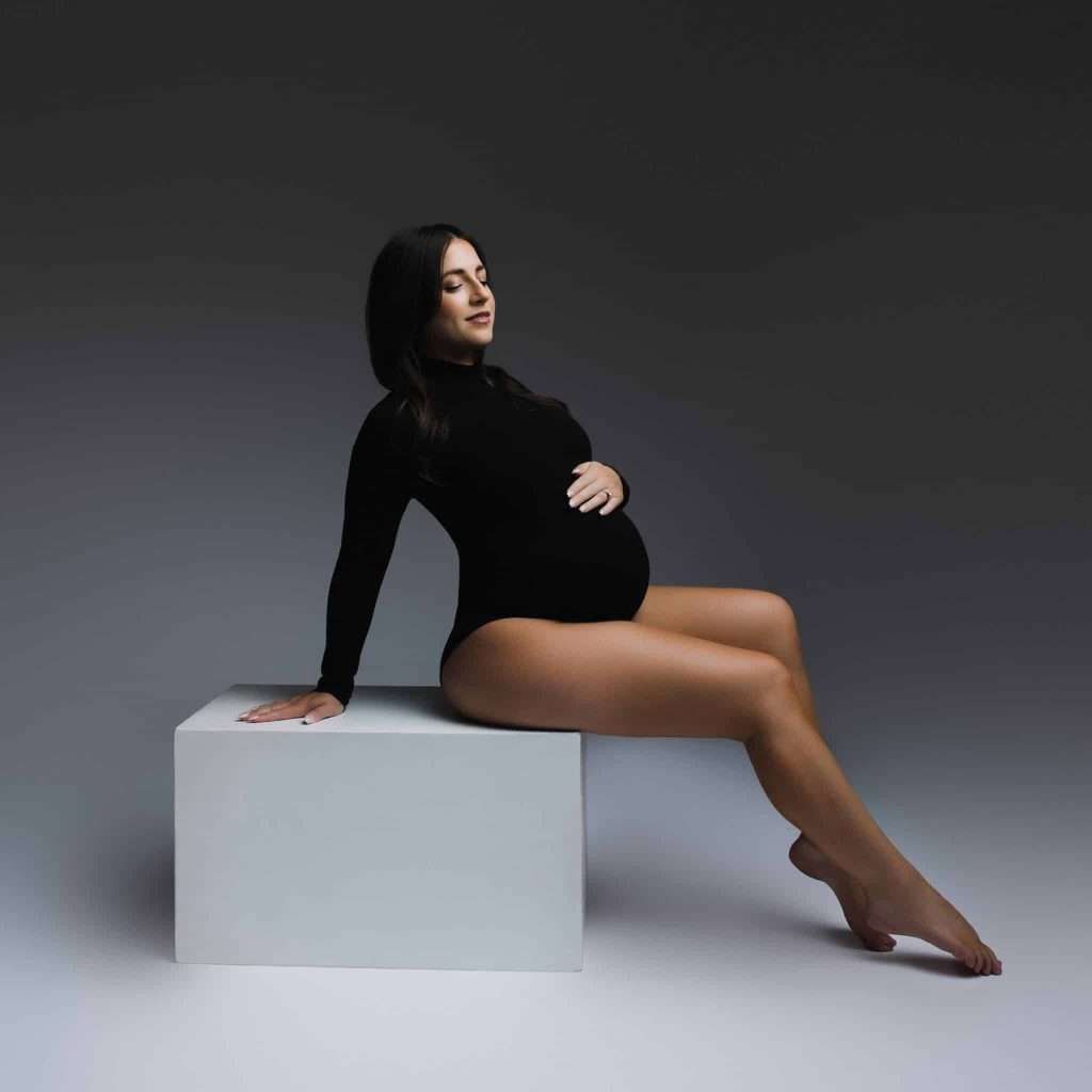 A pregnant woman in a black bodysuit sits on a white block, one hand resting on her knee and the other on her belly, against a plain gray background.