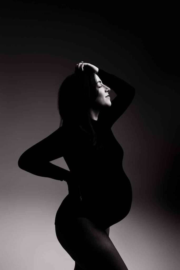 Black and white photo of a pregnant woman in a bodysuit, standing in profile with one hand on her lower back and the other on her head, against a gradient background.
