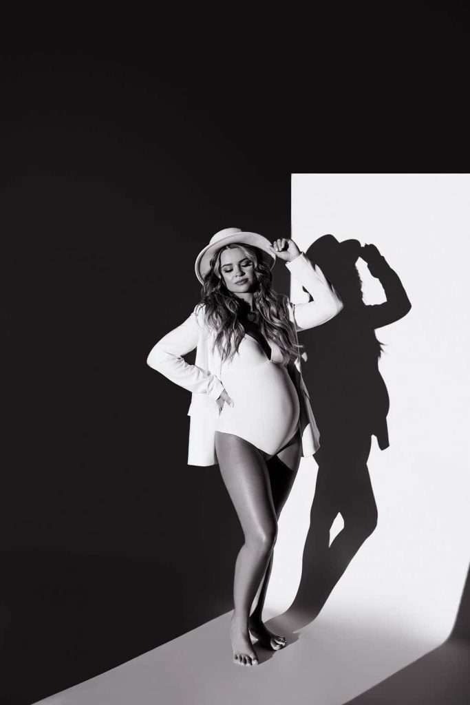 A pregnant woman in a white blazer, bodysuit, and hat stands barefoot, casting a shadow on a black and white background.