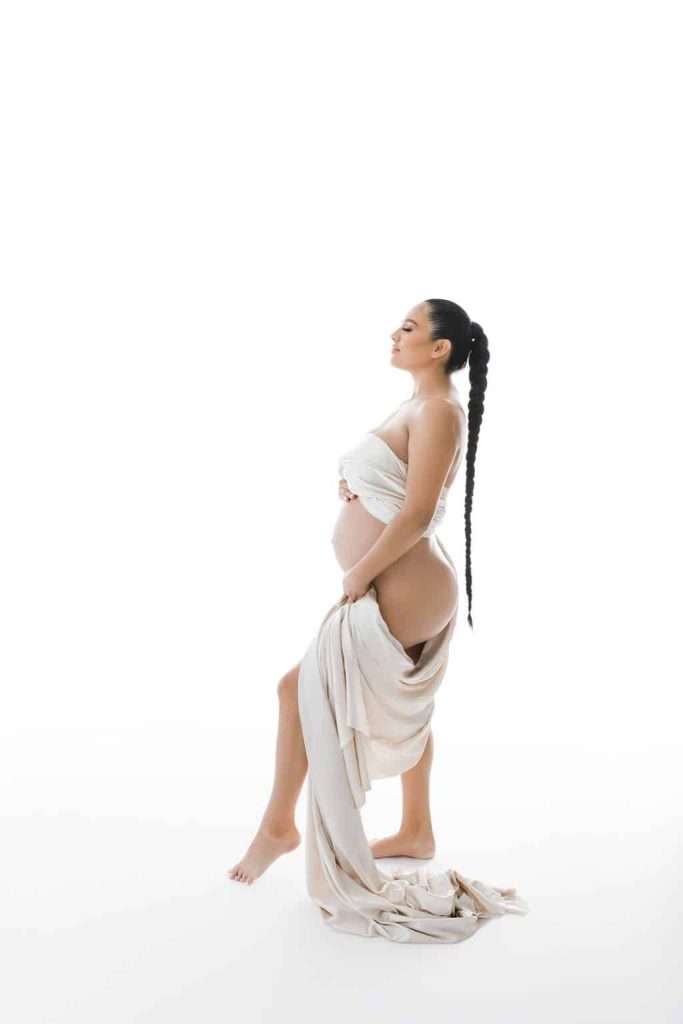 Pregnant woman with a long braid stands in profile, draped in a white cloth, posing against a plain white background.