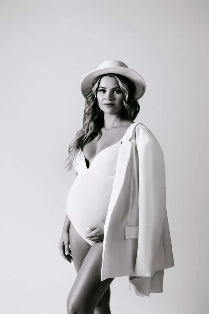 A pregnant woman poses in a bodysuit, jacket draped over one shoulder, and a hat, looking at the camera against a plain background.