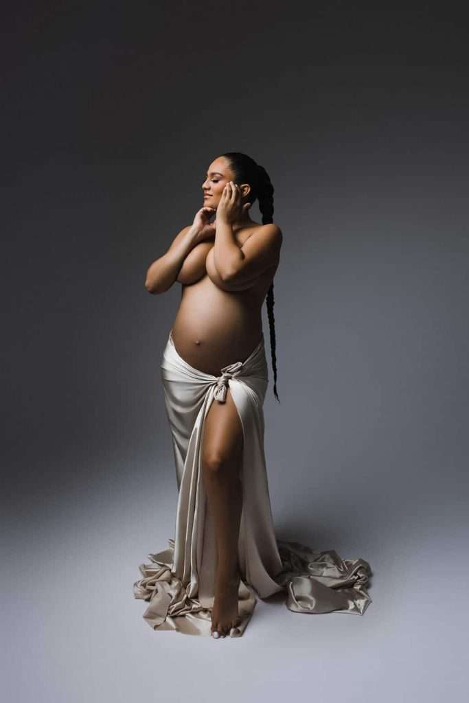 Pregnant woman standing barefoot, draped in a white cloth, with long braided hair, posing against a neutral background.