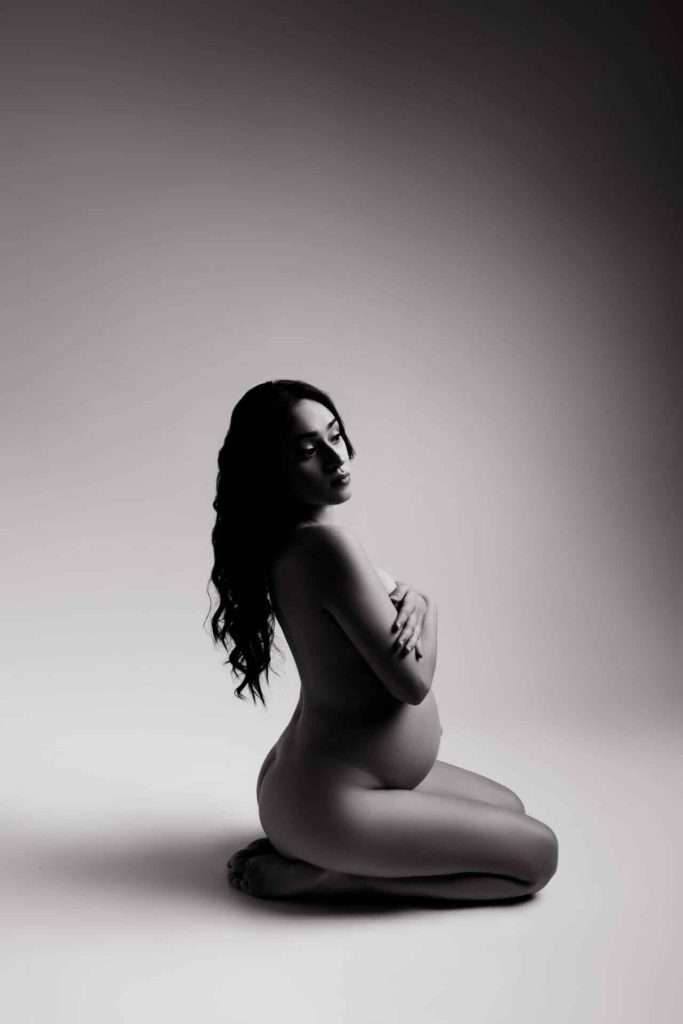 A pregnant woman kneels on the floor, nude, with her arms crossed over her chest in a softly lit studio setting.