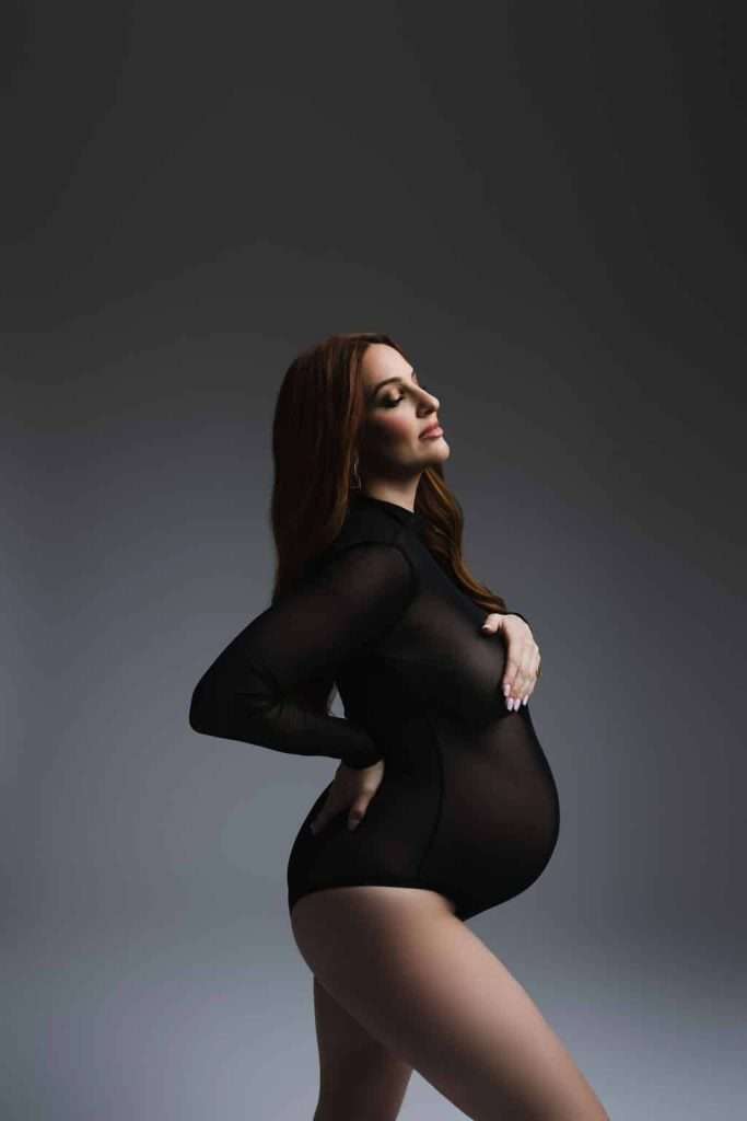 Pregnant woman in a black sheer bodysuit stands in profile, one hand on her lower back and the other supporting her belly, against a neutral gradient background.