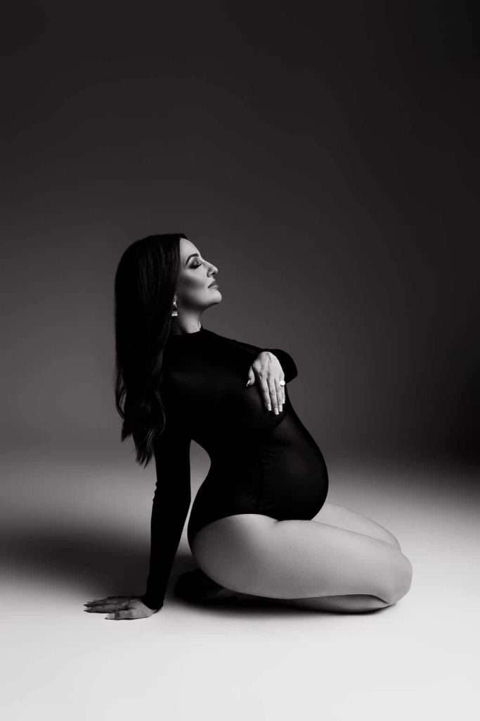 Pregnant woman in a black bodysuit kneeling on the floor, one hand on her belly and the other on the ground, in a studio with dramatic lighting.