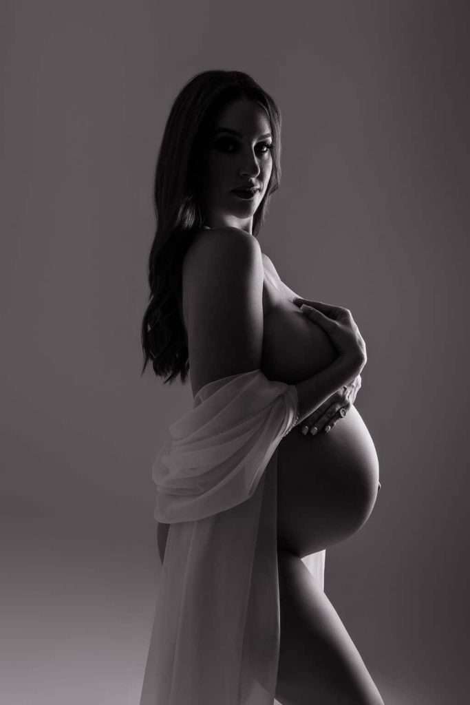 Black and white photo of a pregnant woman in profile, draped in sheer fabric, holding her belly and looking at the camera.