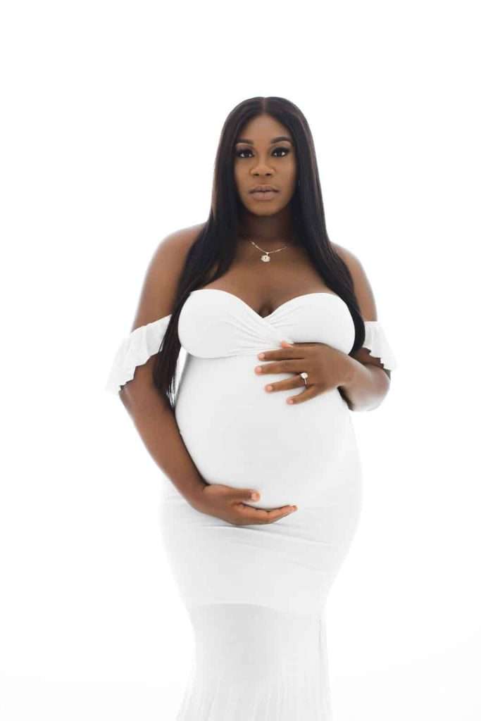 A pregnant woman in a fitted white dress stands against a white background, holding her belly with both hands and looking at the camera.
