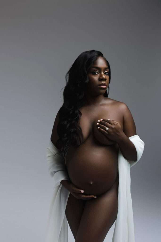 Pregnant woman with long hair poses nude, partially draped in a sheer white fabric, holding her belly and covering her chest, against a neutral background.