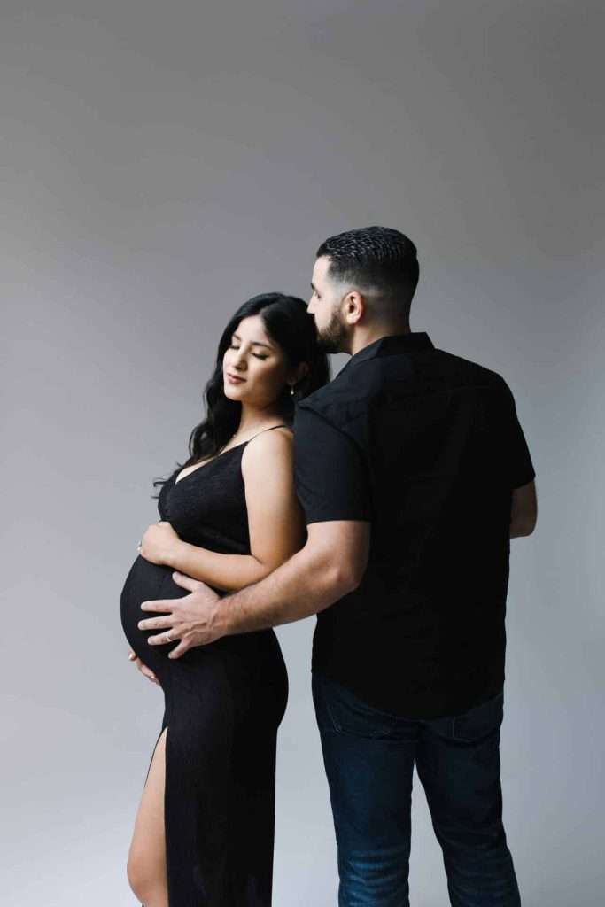 A man stands behind a pregnant woman in a black dress, gently holding her belly while she closes her eyes. Both face sideways against a plain background.