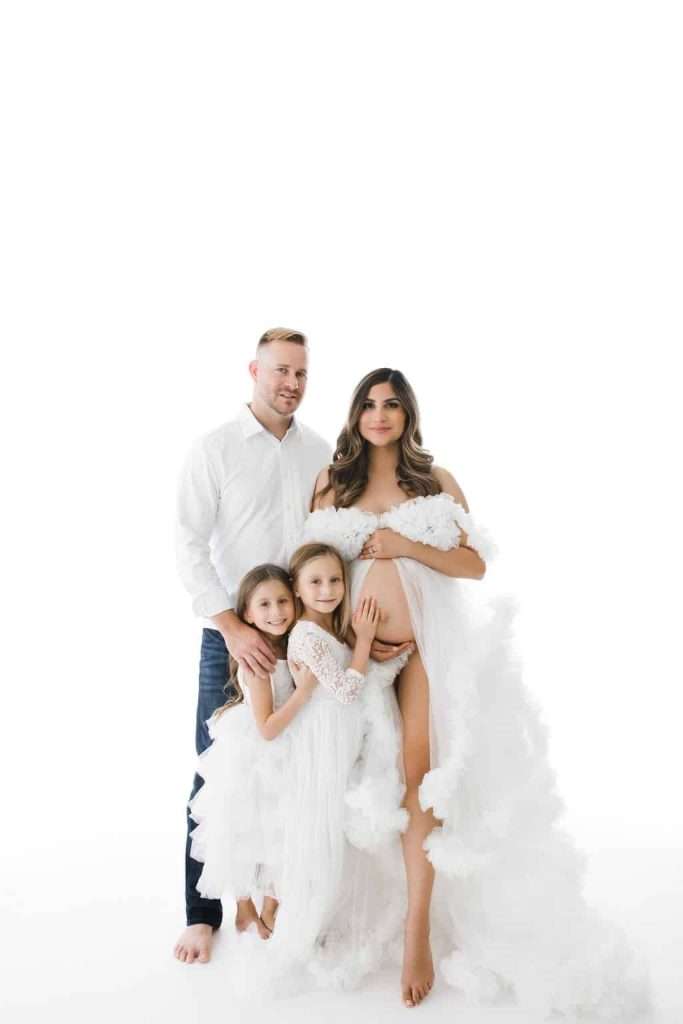 A family of four poses in white outfits against a white background; the mother is visibly pregnant, and two young girls stand in front.