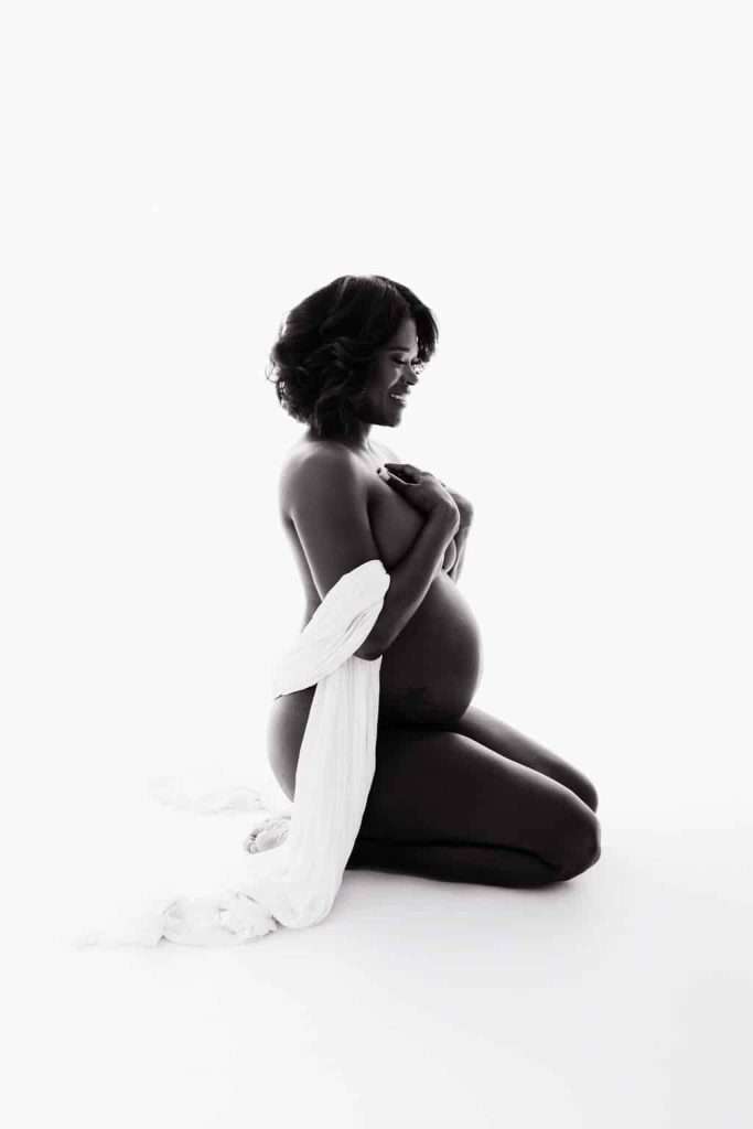 A pregnant woman kneels on the floor, covering her chest with her hands and draped in a white cloth, against a plain white background.
