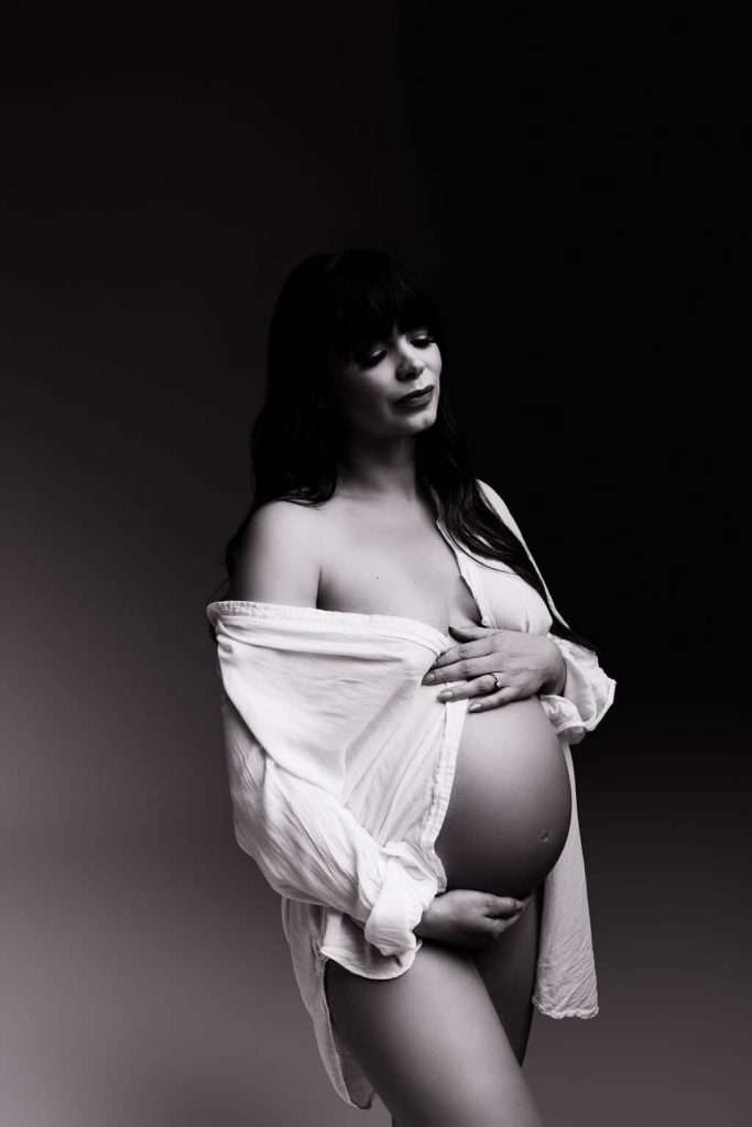 A pregnant woman in a partially open white shirt stands against a dark background, holding her belly with one hand and covering her chest with the other.