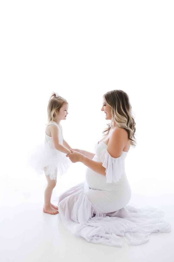 Pregnant woman kneels on the floor in a white dress, holding hands with a smiling young girl in a white dress, against a bright white background.