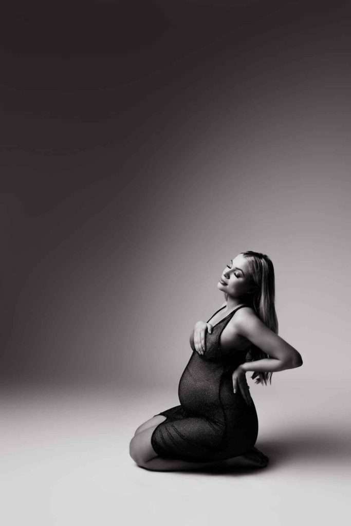 Pregnant woman in a black dress kneels on the floor, one arm on her belly and the other behind her back, with eyes closed in a softly lit studio.