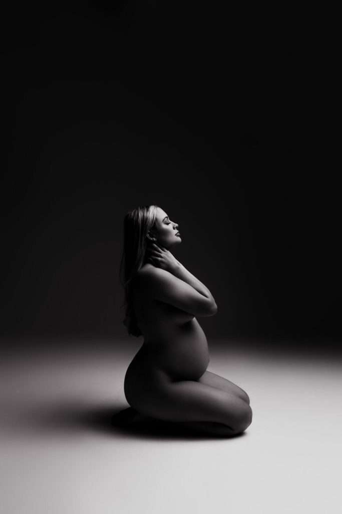 A pregnant woman kneels on the ground in a dimly lit studio, holding her neck and looking upward, with dramatic lighting highlighting her silhouette.