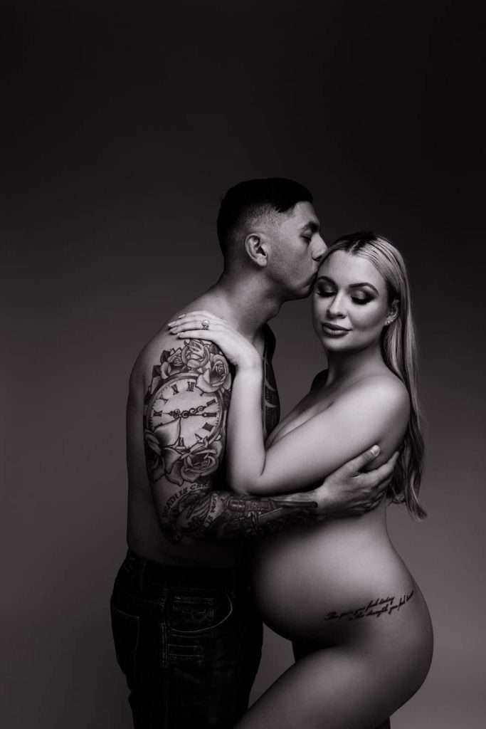 A shirtless man with tattoos kisses the forehead of a pregnant woman, both standing and embracing against a plain background.
