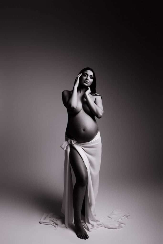 A pregnant woman stands barefoot, draped in a white cloth that covers her lower body, posing in a studio with a neutral background.