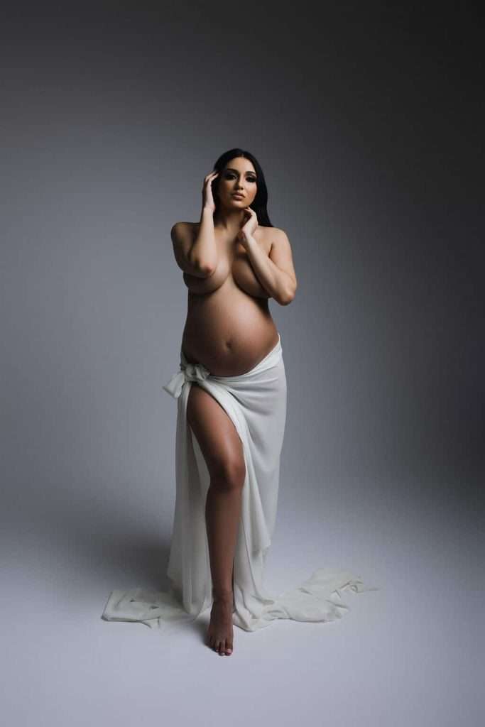 Pregnant woman poses in studio, standing with one knee raised, draped in a white cloth that covers her lower body, against a neutral gray background.