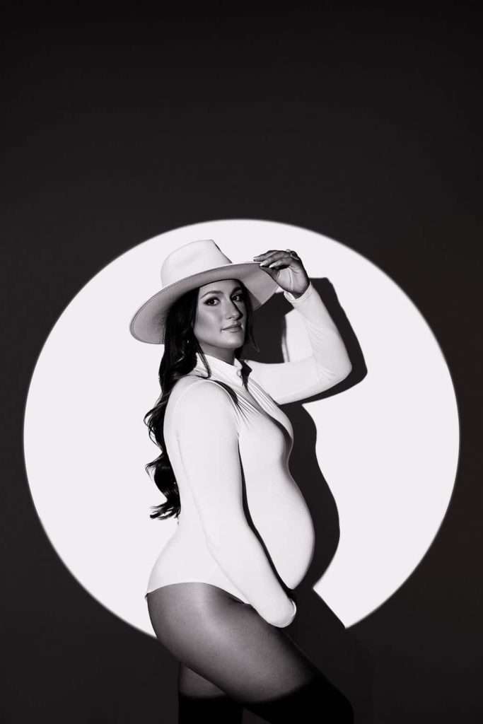 Pregnant woman in a white bodysuit and hat poses in front of a circular light backdrop, looking at the camera.