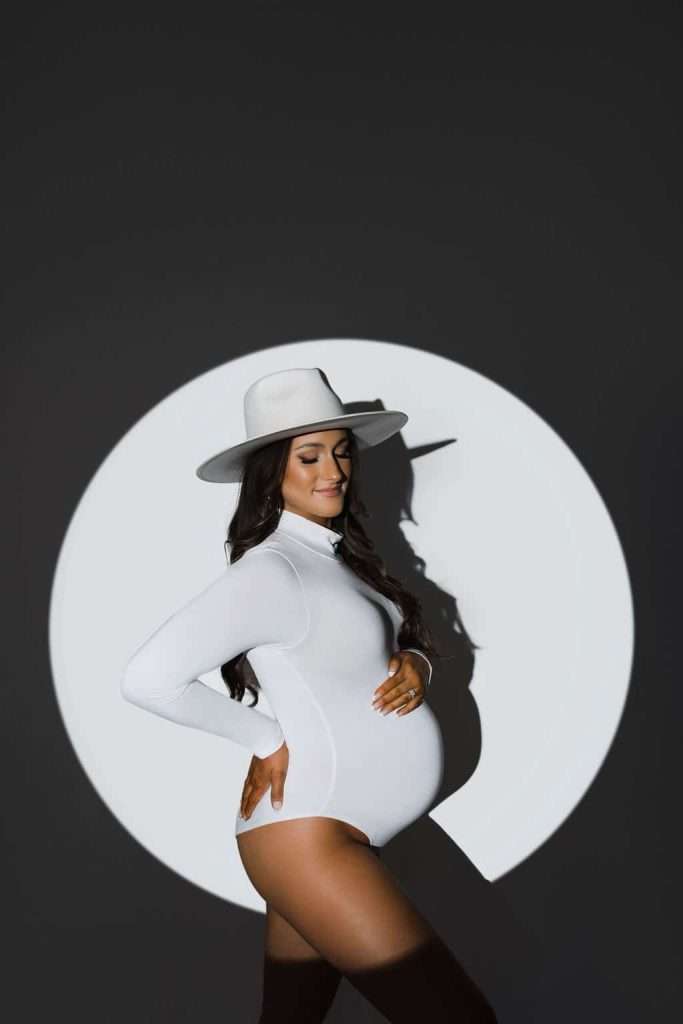 Pregnant woman in a white bodysuit and wide-brimmed hat poses in front of a dark background with a white circle of light.