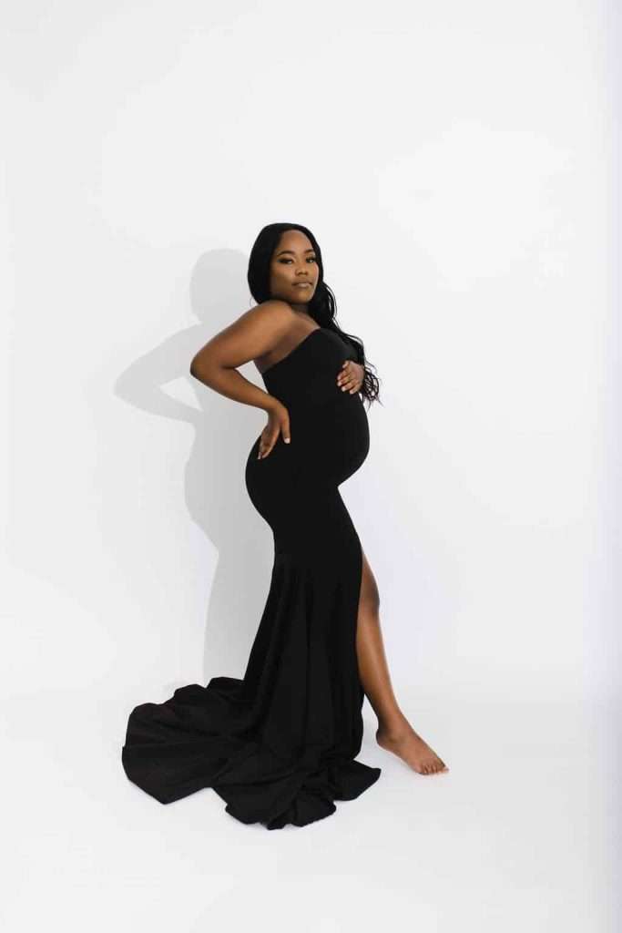 Pregnant woman in a black, floor-length gown poses barefoot against a plain white background, with one hand on her hip and the other on her belly.