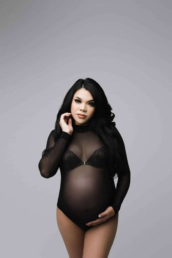 Pregnant woman in a black sheer bodysuit stands against a plain gray background, one hand on her belly and the other touching her hair.